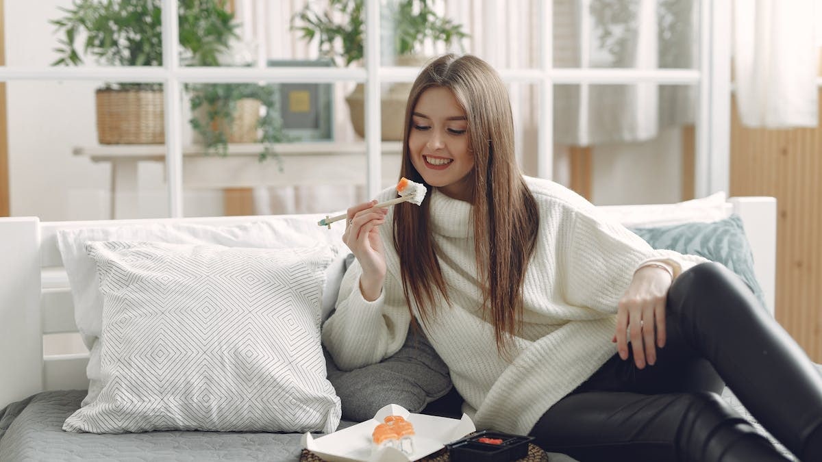 Smiling young lady in warm knitted sweater enjoying fresh sushi on wicker tray while relaxing on cozy sofa with soft cushions in modern living room